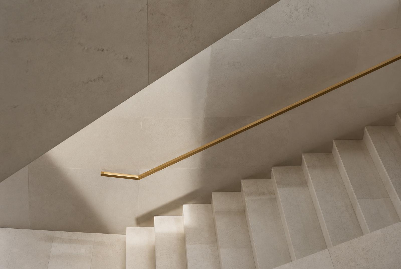 Pale stone stairs seen from above with clean diagonals, soft shadow and a slim brass handrail.